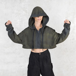 Person facing forward from head to knees with their arms slightly raised wearing a green and black patterned cropped hoodie with the hood up to cover their face against a light gray background