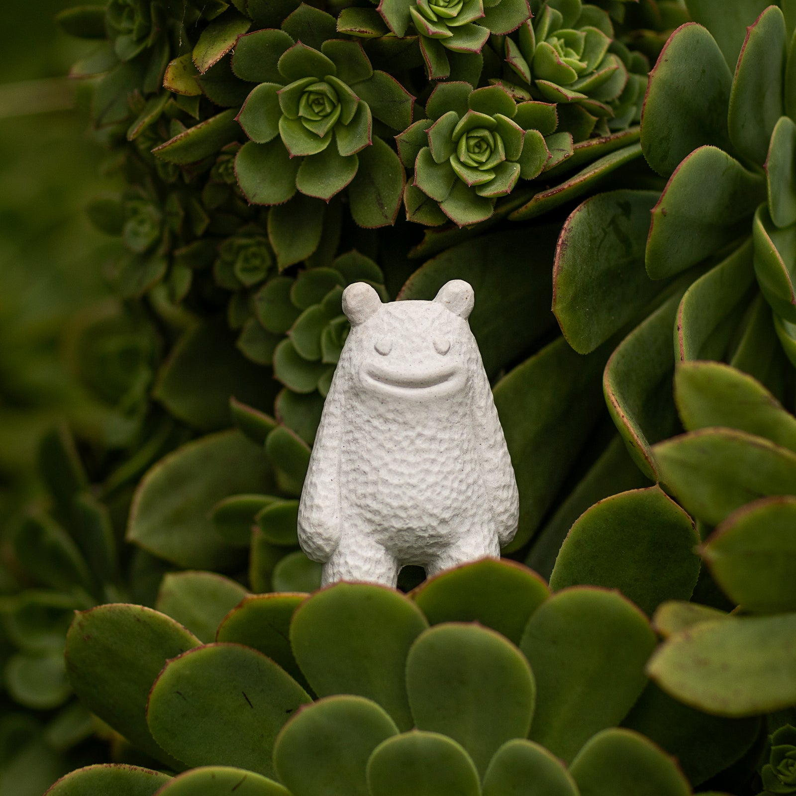 Small white ceramic smiling creature figurine standing on green succulents 