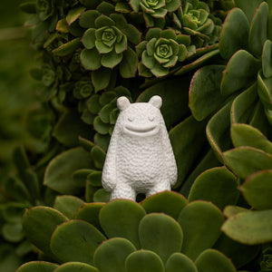 Small white ceramic smiling creature figurine standing on green succulents 