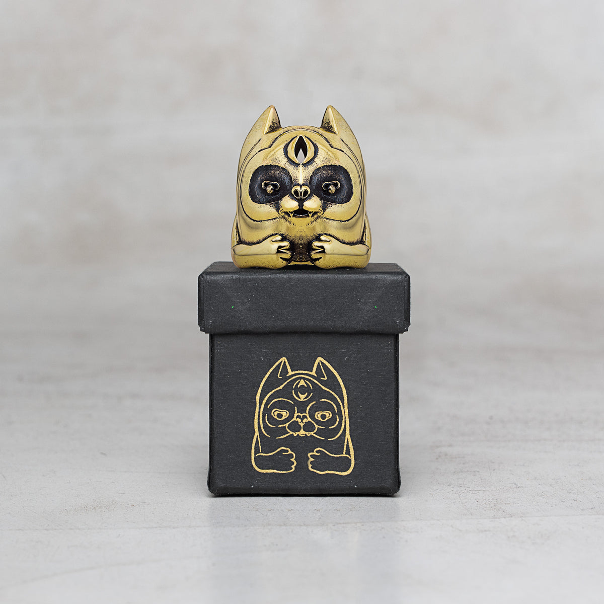 A small gold cat figurine with black facial details sits on top of a black box featuring a gold cat illustration, photographed against a light gray background.