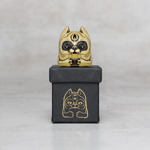 A small gold cat figurine with black facial details sits on top of a black box featuring a gold cat illustration, photographed against a light gray background.