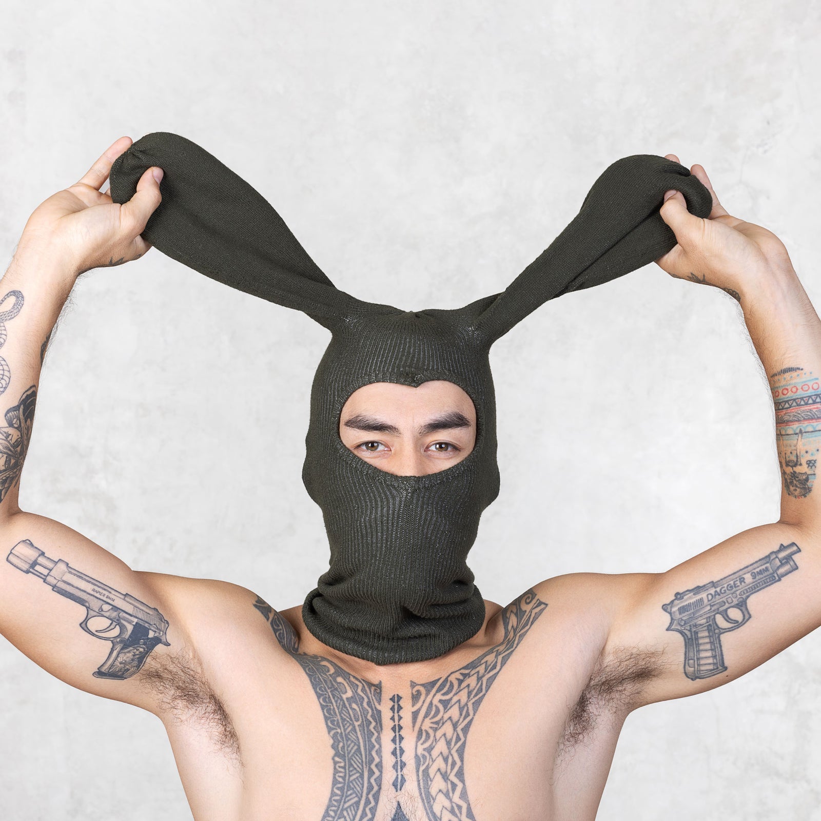 Person wearing a dark olive bunny mask with ears, arms outstretched, on a light gray background.