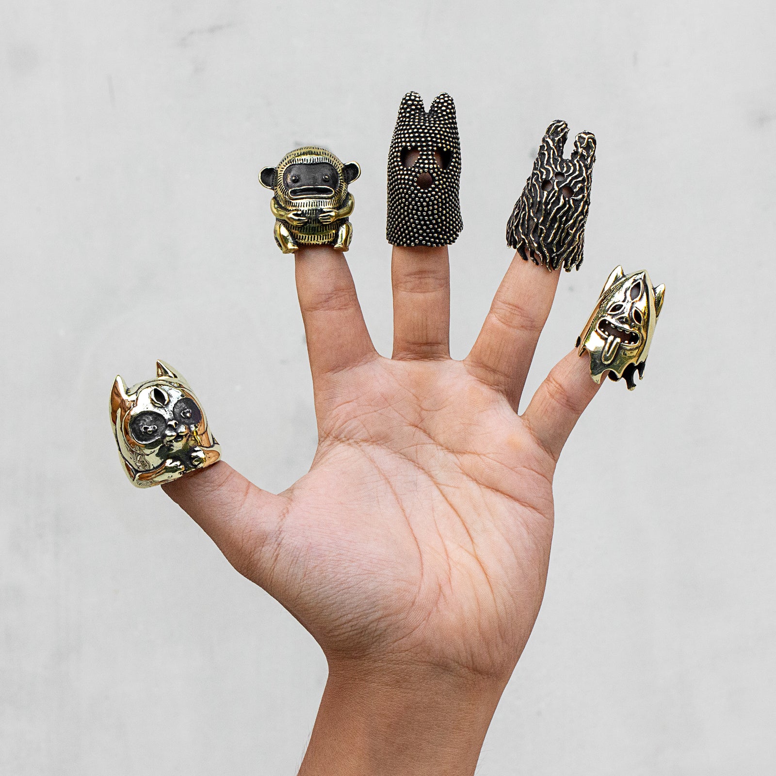 A hand displaying five unique finger puppets against a light gray background. Crafted in gold brass with black accents.