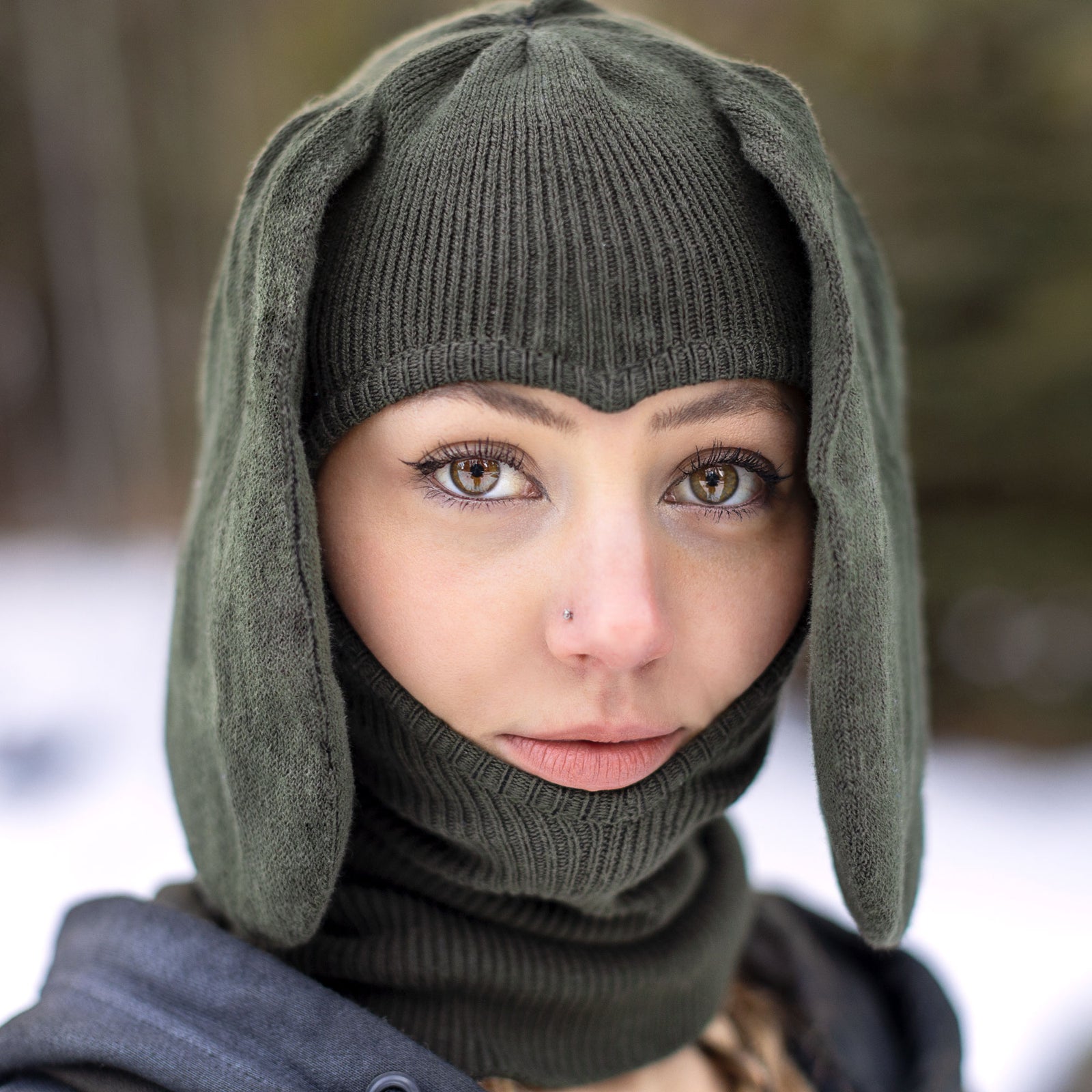 Person wearing a green knitted beanie balaclava hat with bunny ears and e heart shaped face hole outdoors.