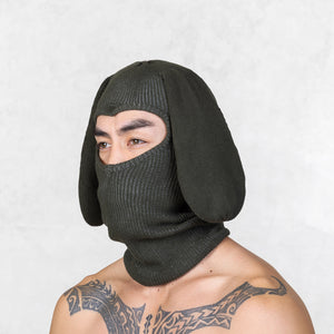 Person wearing a dark green knit balaclava with floppy bunny ears on a light gray background