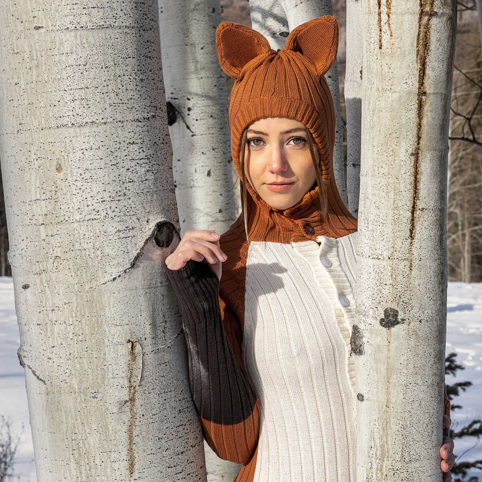 Person wearing a fox sweater with hooded ears between trees in a snowy forest 