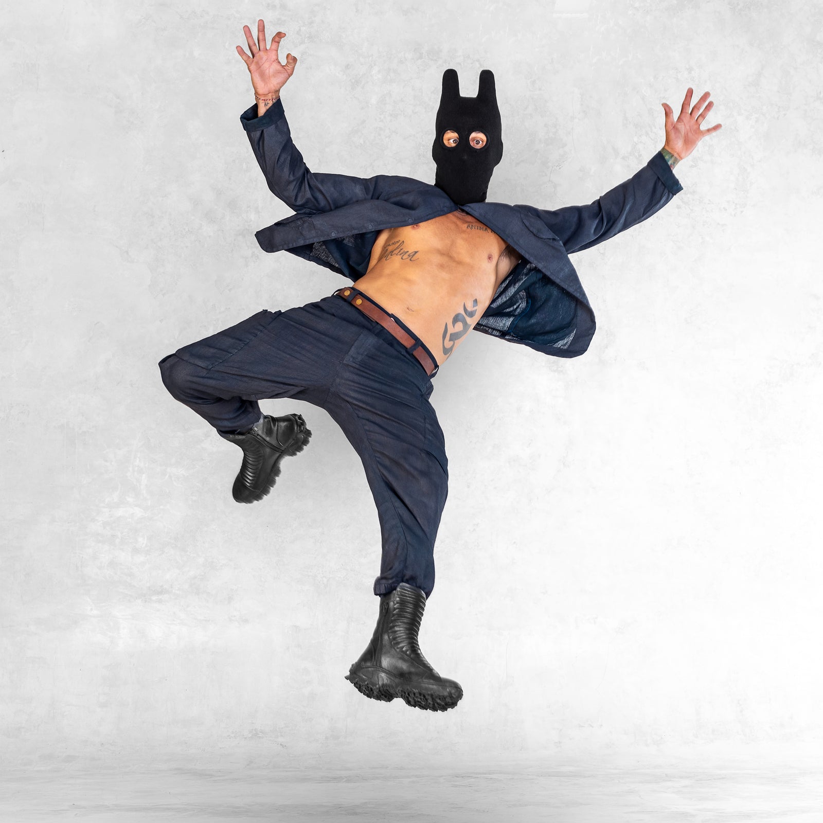 Person wearing a blue linen shirt and pants, a black balaclava mask, a brown belt with brass accents, and black leather boots jumping with arms outstretched against a light gray background