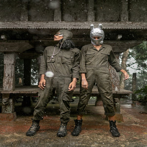 Two individuals in green military-style uniforms standing under a wooden structure.