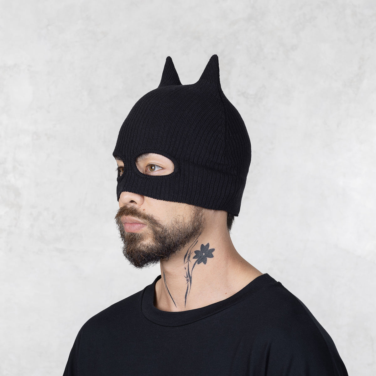 Person wearing a black knit bat mask with ears on a light gray background