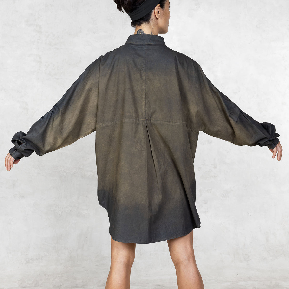 Person facing back wearing hand-painted olive tunic button up shirt with arms extended against a white background