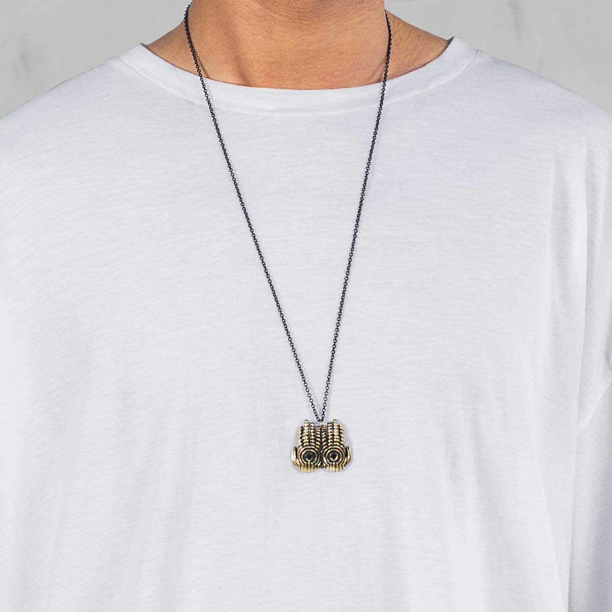 A close-up of a person wearing a white t-shirt and a long black chain necklace with a textured gold pendant.