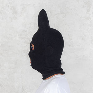 Person facing sideways wearing a black balaclava with bat ears against a light gray background