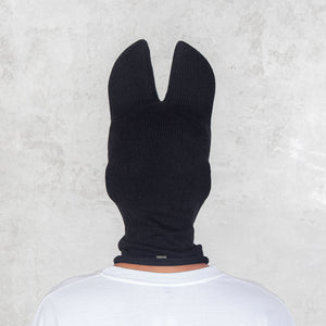 Person facing backwards wearing a black balaclava with bat ears against a light gray background