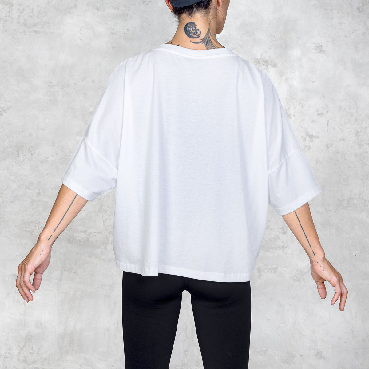 Tattooed person facing back wearing a white oversized t-shirt and black pants against a white background