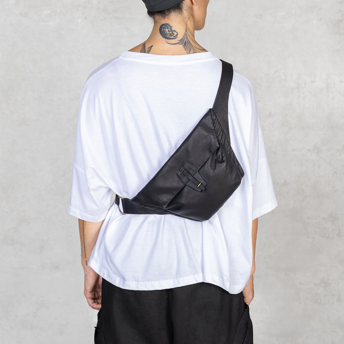 Person wearing a white t-shirt, black pants, and a black sling bag over one shoulder facing backwards against a plain background