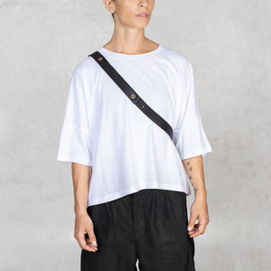 Person wearing a white t-shirt, black pants, and a black strap over one shoulder against a plain background