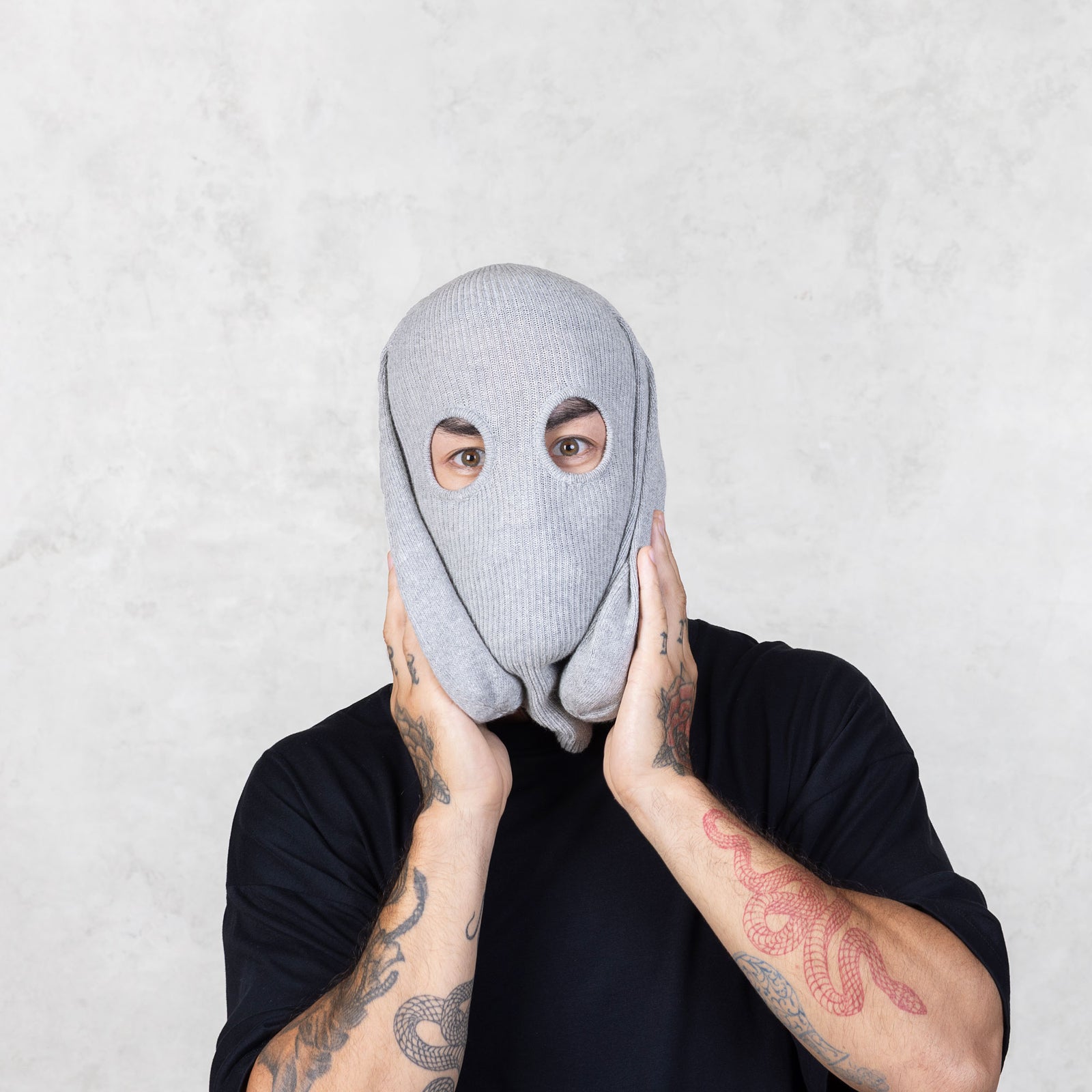 Person with tattooed arms wearing a gray knit bunny balaclava holding the ears down against a plain background