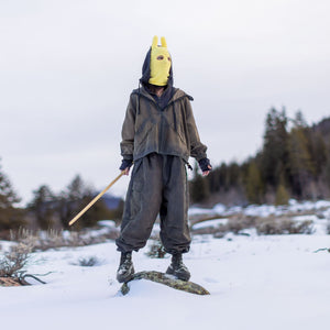 Person in a yellow mask and dark outfit standing in a snowy landscape with trees in the background.