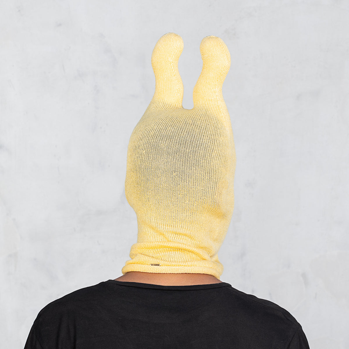 A person viewed from the back wearing a pale yellow knit balaclava-style hood with two upright ears, paired with a black shirt, against a light gray marble-textured background.