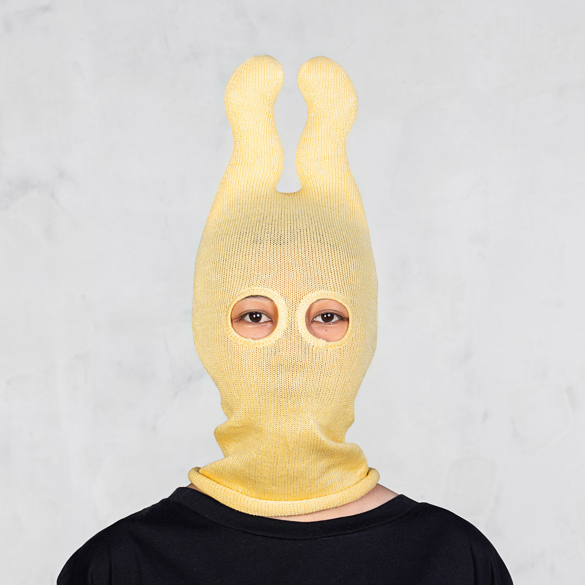 A person wearing a pale yellow ribbed knit balaclava-style hood with two upright bunny ears and eye cutouts, shown against a light gray wall.