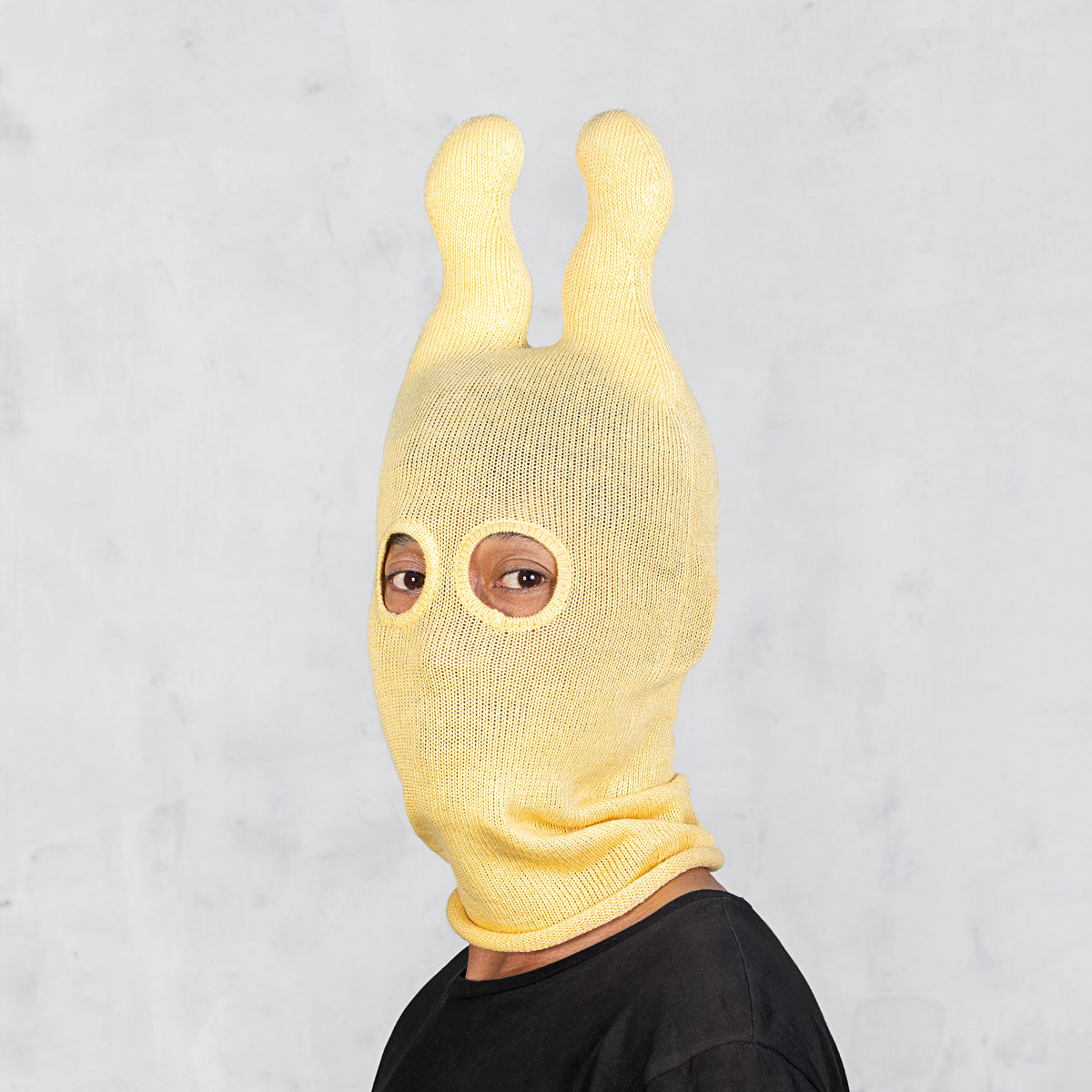 A person wearing a yellow knit balaclava-style hood with two upright wavy ears and eye cutouts, photographed against a light gray wall.