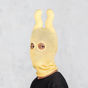 A person wearing a yellow knit balaclava-style hood with two upright wavy ears and eye cutouts, photographed against a light gray wall.