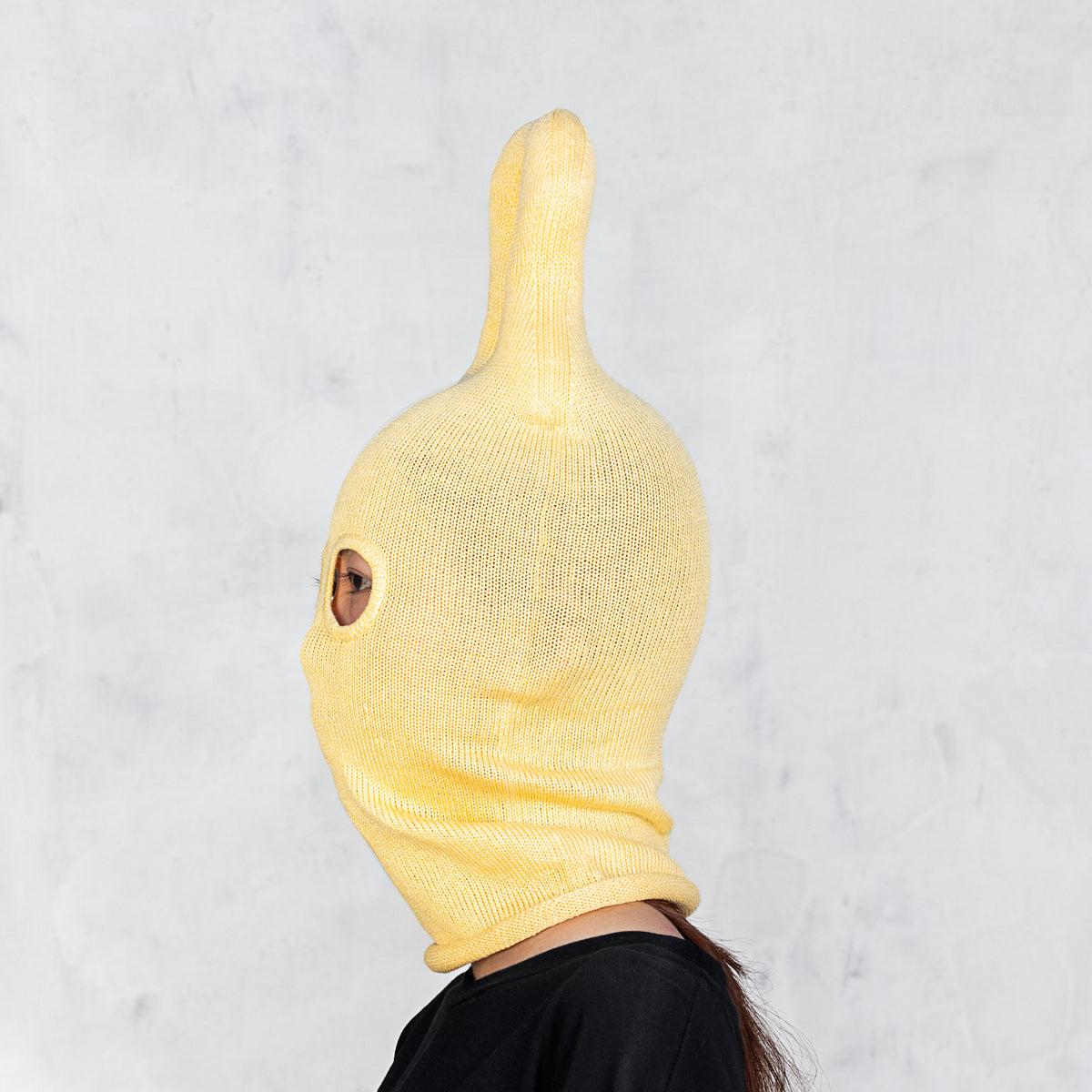 A person wearing a yellow ribbed knit balaclava-style hood with upright horn-like protrusions on top, shown in side profile against a light gray textured wall.