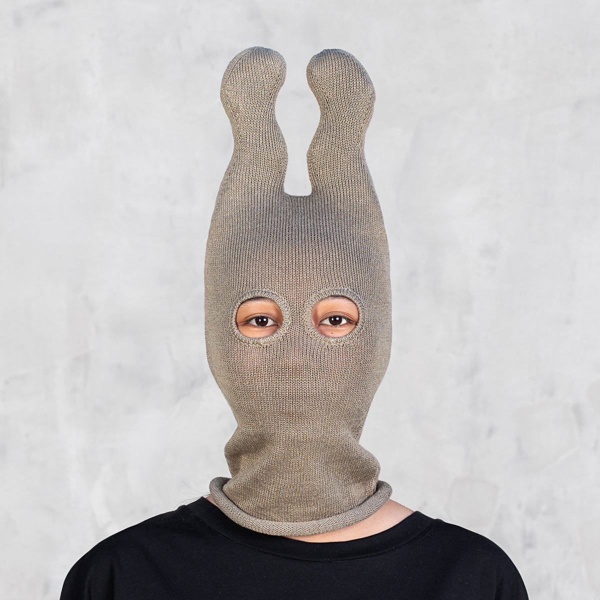 A person wearing an olive knit balaclava-style mask with two upright wavy ears, shown from the shoulders up against a light gray textured wall.