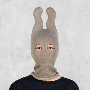 A person wearing an olive knit balaclava-style mask with two upright wavy ears, shown from the shoulders up against a light gray textured wall.