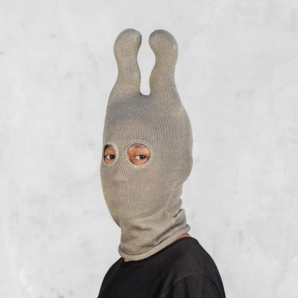 A person wearing an olive ribbed knit balaclava with two wavy bunny ears and eye cutouts, shown against a light textured wall.