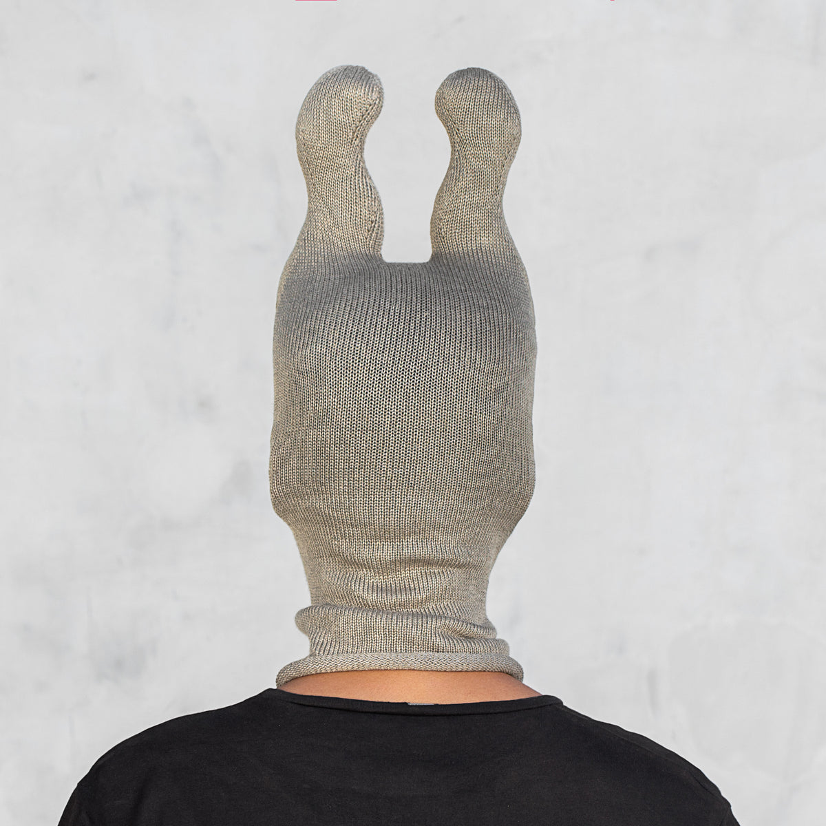 A person shown from the back wearing a ribbed knit balaclava-style head covering with two upright bunny ears, in an olive color over a black shirt, against a light gray wall.