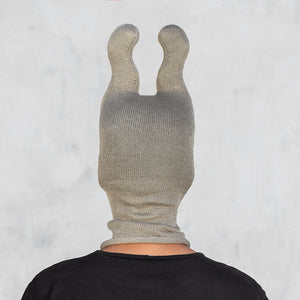A person shown from the back wearing a ribbed knit balaclava-style head covering with two upright bunny ears, in an olive color over a black shirt, against a light gray wall.