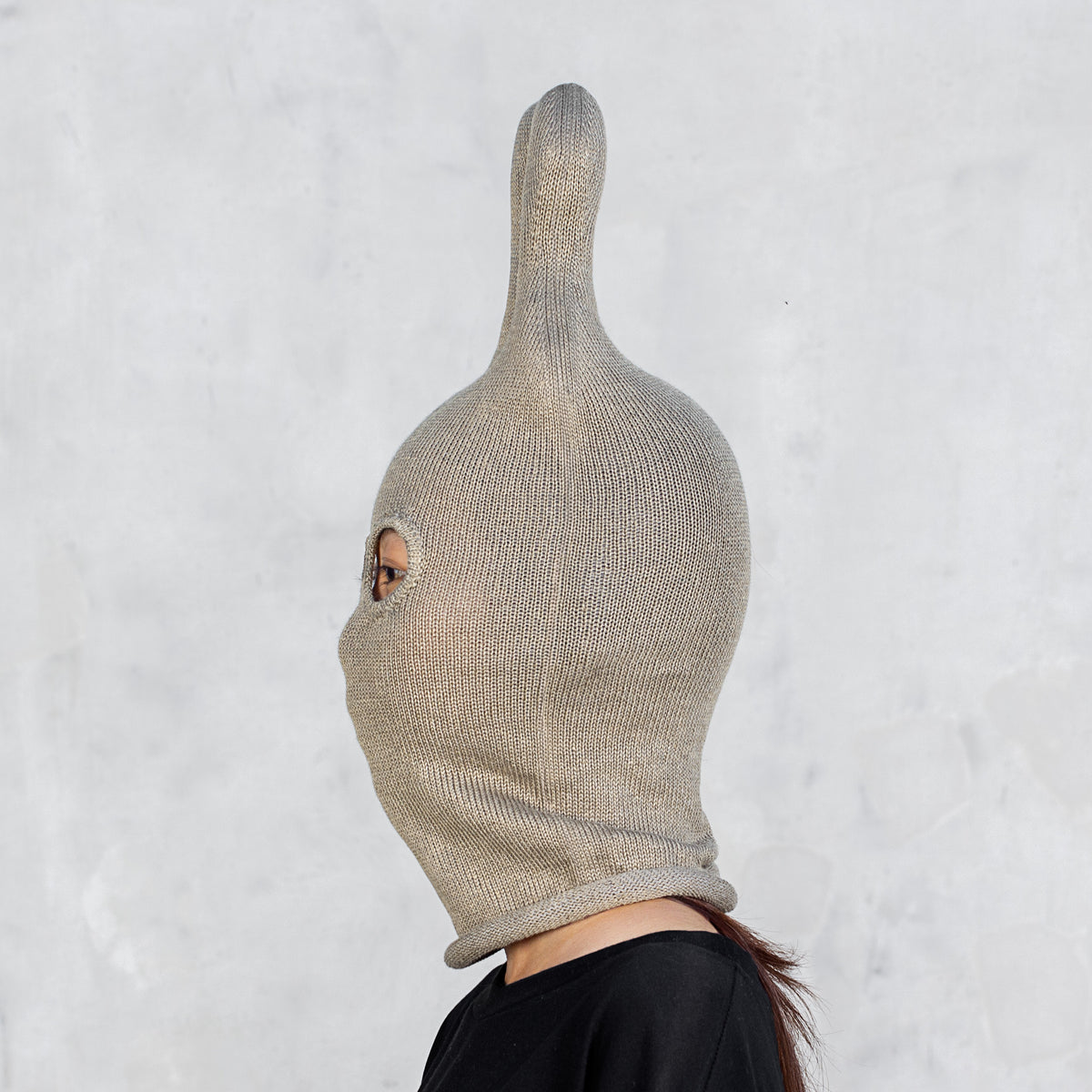 A person shown in side profile wearing an olive ribbed knit hood with a tall pointed top and a small eye opening, against a light gray textured wall.