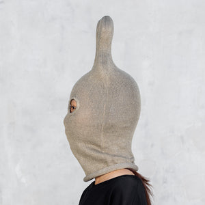 A person shown in side profile wearing an olive ribbed knit hood with a tall pointed top and a small eye opening, against a light gray textured wall.