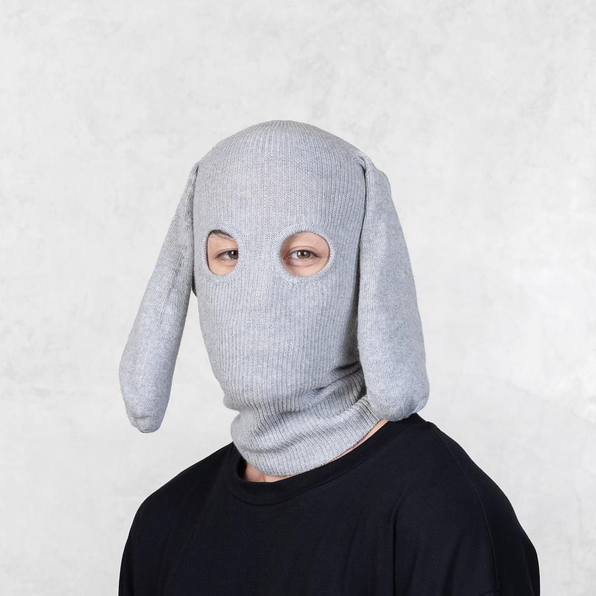 Person wearing a gray knit balaclava with floppy ears against a light gray background 