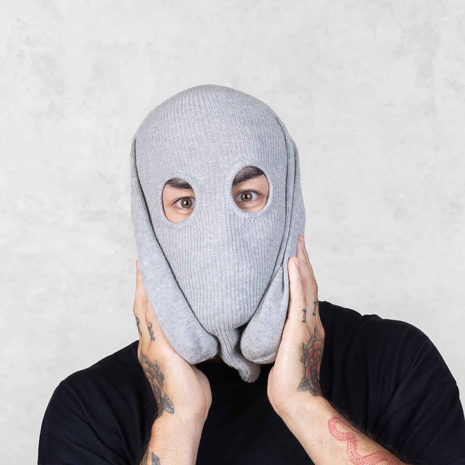 Person wearing a gray knit balaclava holding the floppy ears to their face against a light gray background 