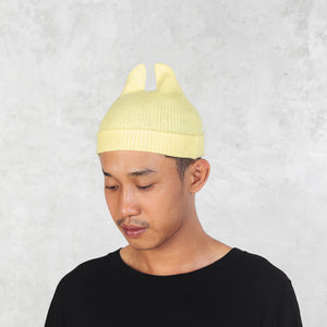 Person looking down wearing a light yellow beanie with spikey ears against a plain background