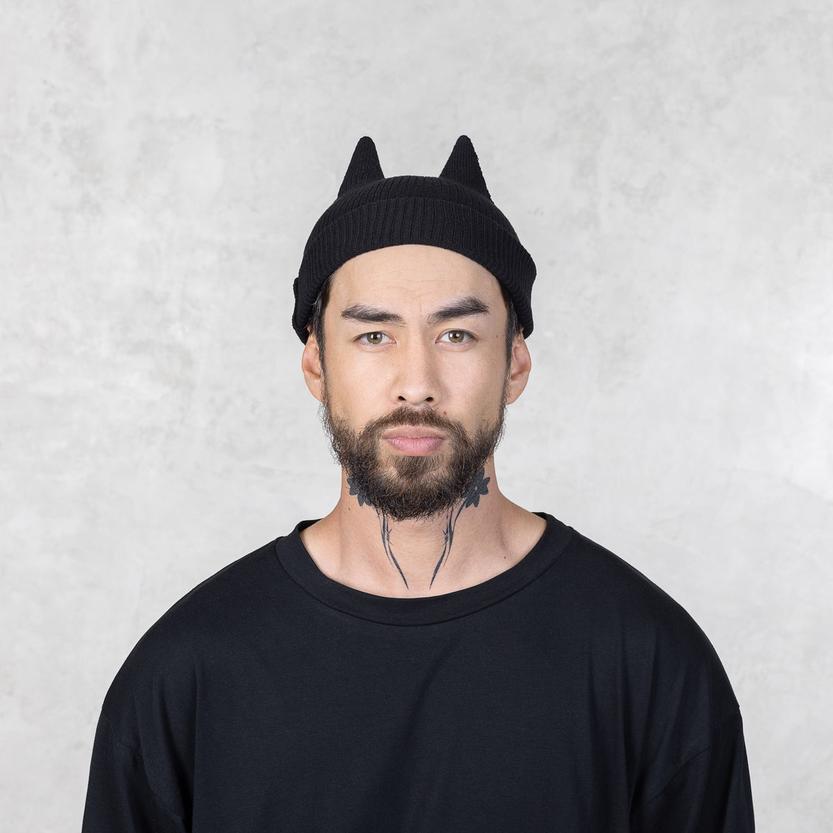 Person with a mustache and beard wearing a black beanie with spike ears against a light gray background