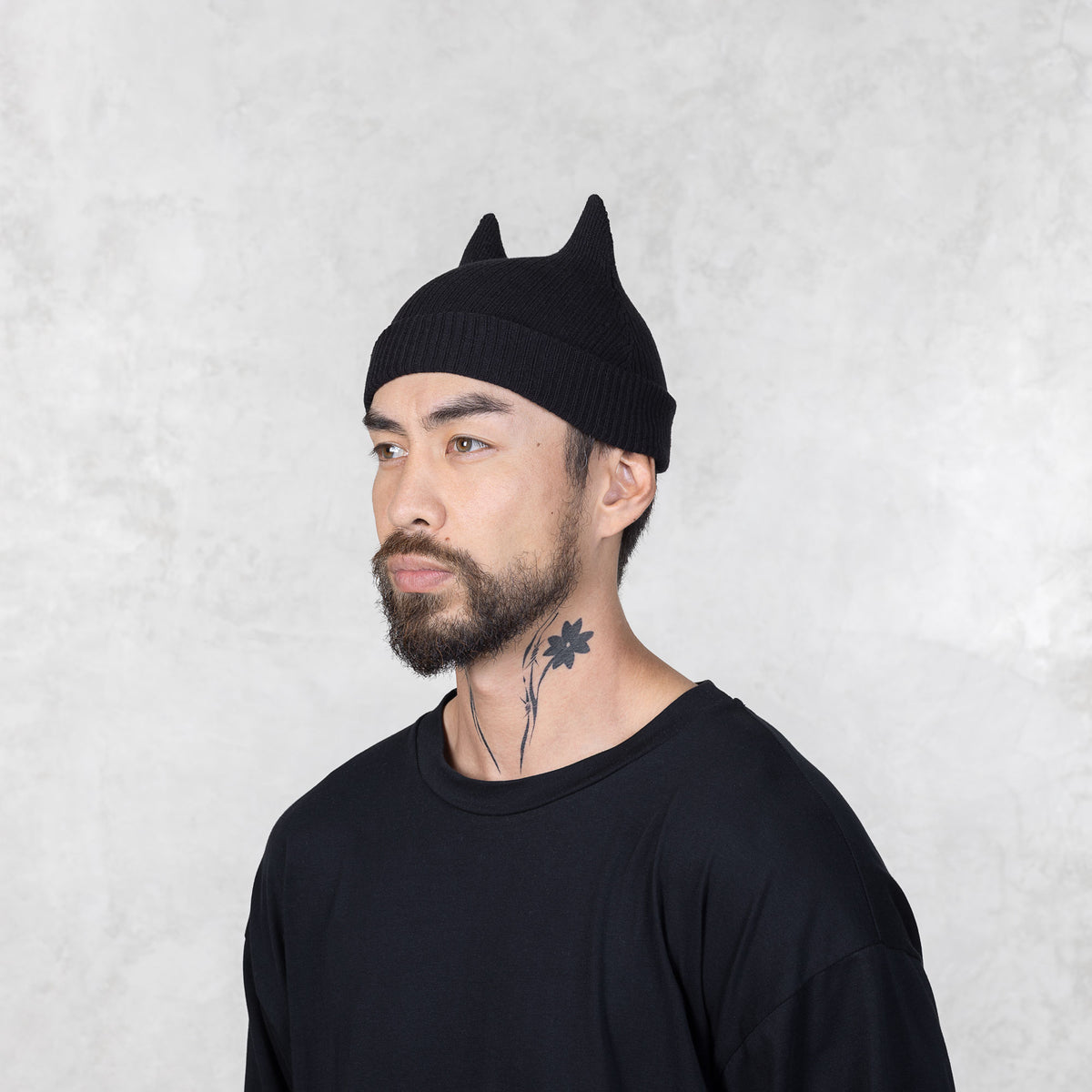 Person with a mustache and beard looking away wearing a black beanie with spike ears against a light gray background