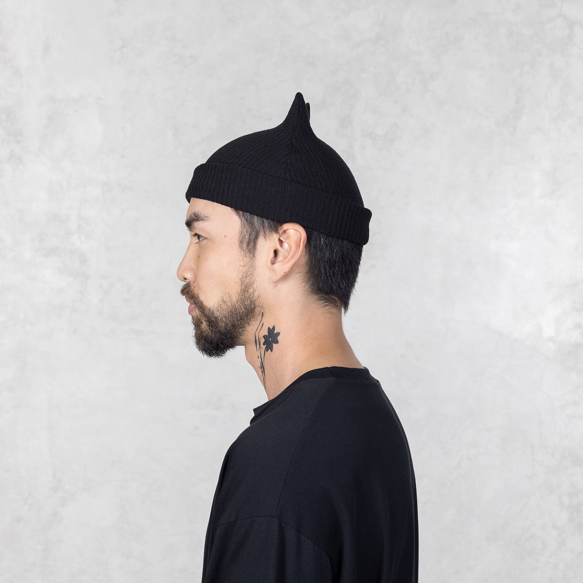 Person with a mustache and beard facing sideways wearing a black beanie with spike ears against a light gray background