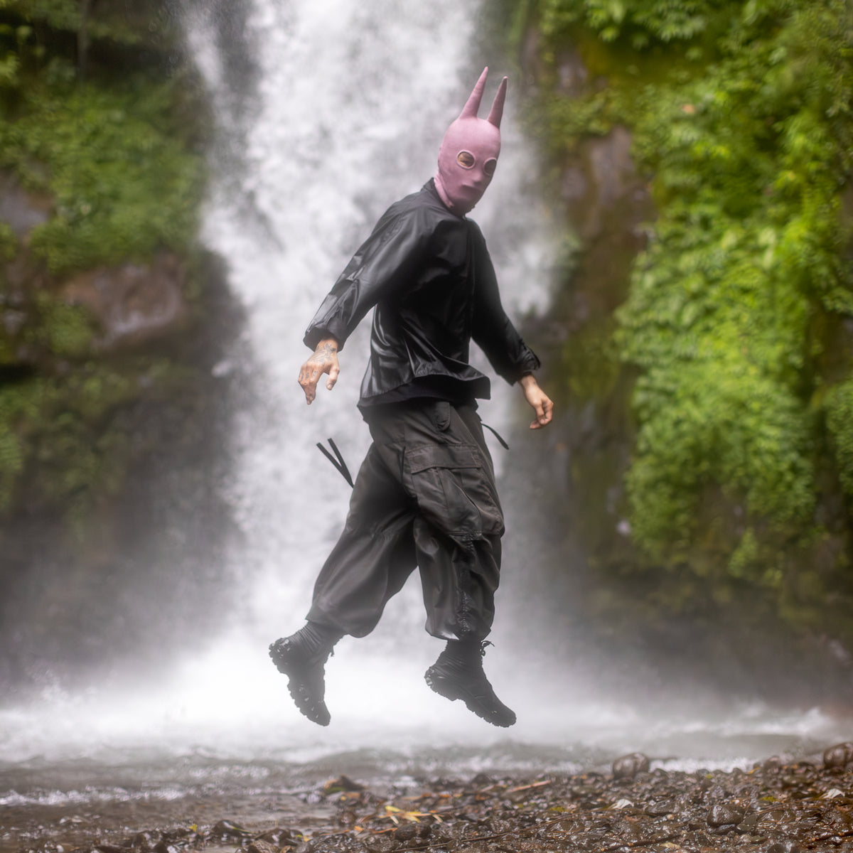 A person in an olive jumpsuit with a Light pink Spire balaclava in front of a waterfall
