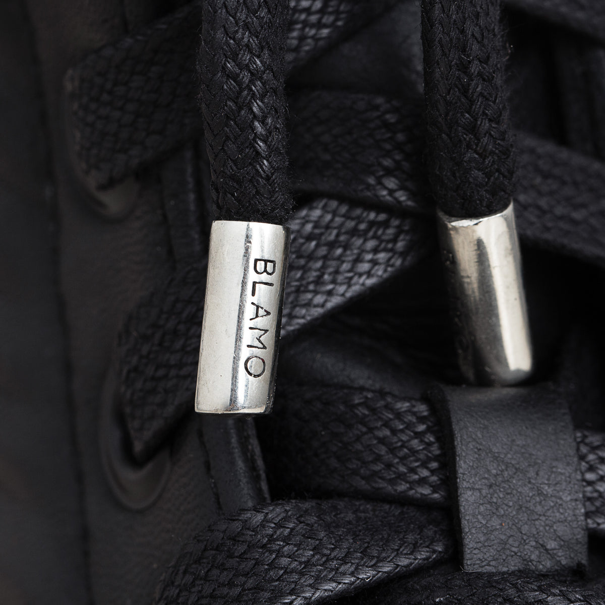 Close-up of black laces threaded through a black shoe, with metallic silver eyelets and the word 'BLAMO' engraved on one lace tip.