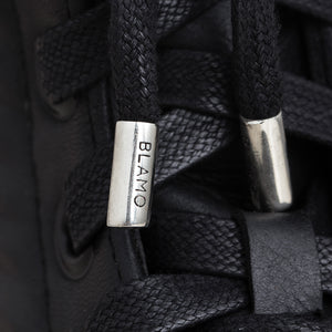 Close-up of black laces threaded through a black shoe, with metallic silver eyelets and the word 'BLAMO' engraved on one lace tip.