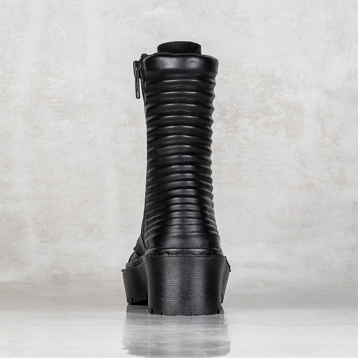 A single black mid-calf boot with a vertically ribbed shaft and a chunky platform sole, shown from the back against a textured light gray background.