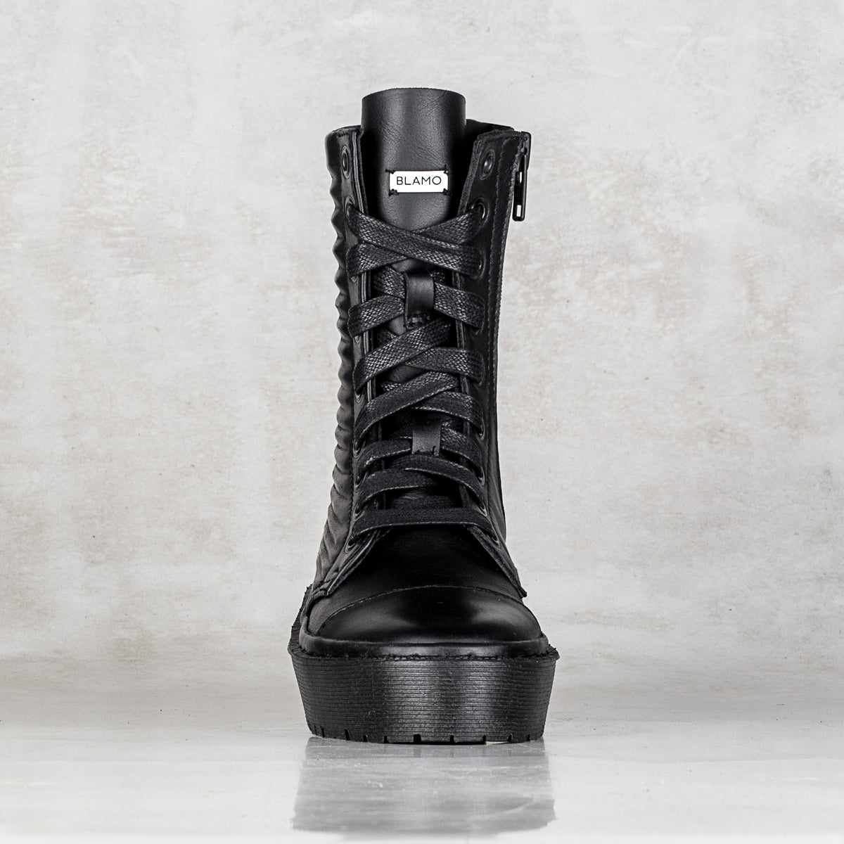 A single black lace-up ankle boot with a thick platform sole, textured side detailing, and a visible 'BLAMO' brand tag near the collar, set against a light gray textured background.