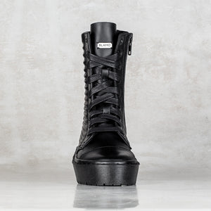 A single black lace-up ankle boot with a thick platform sole, textured side detailing, and a visible 'BLAMO' brand tag near the collar, set against a light gray textured background.