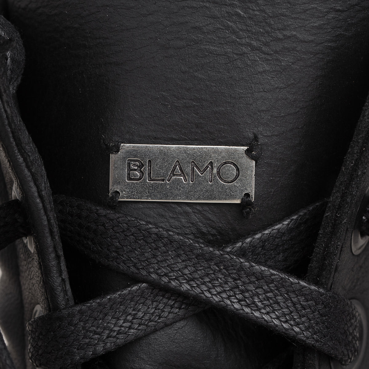 Close-up of a black leather shoe with black laces and a rectangular metal brand plaque engraved with 'BLAMO'.