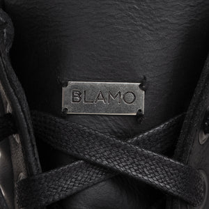 Close-up of a black leather shoe with black laces and a rectangular metal brand plaque engraved with 'BLAMO'.