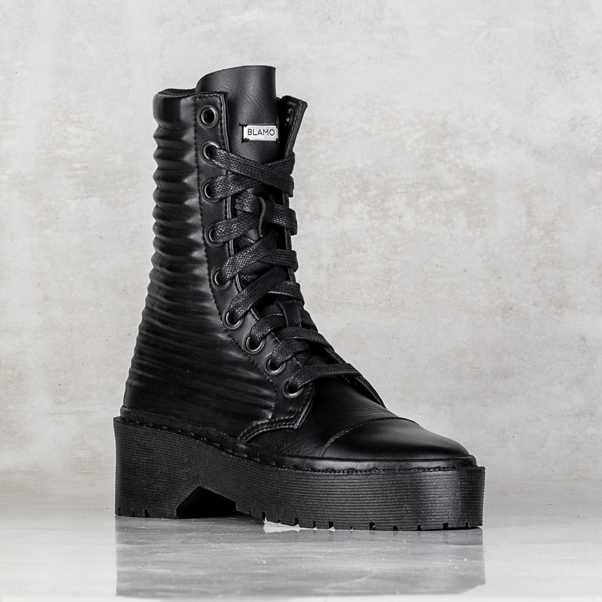 A single black lace-up ankle boot with a ribbed textured shaft, chunky platform sole, and a small silver BLAMO label on the tongue, set against a textured gray background.
