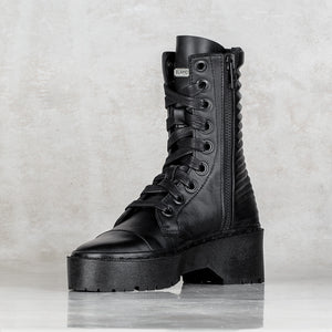 A single black leather lace-up ankle boot with a thick sole, side zipper, and decorative ridged heel detail, shown against a light gray textured background.